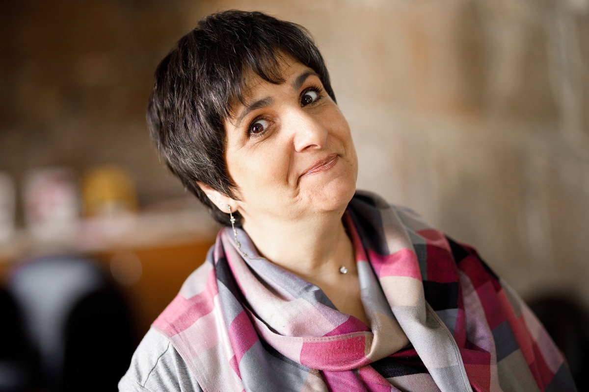 kobieta w wieku 40 plus z krótkimi ciemnymi włosami i w szalu stoi z kpiącym uśmiechem