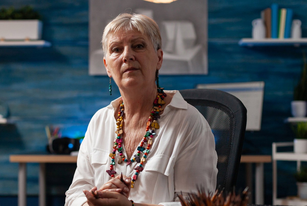 kobieta w wieku 55 plus z krótkimi włosami i koralami na szyi siedzi z ołówkiem w ręku smutna i zamyślona