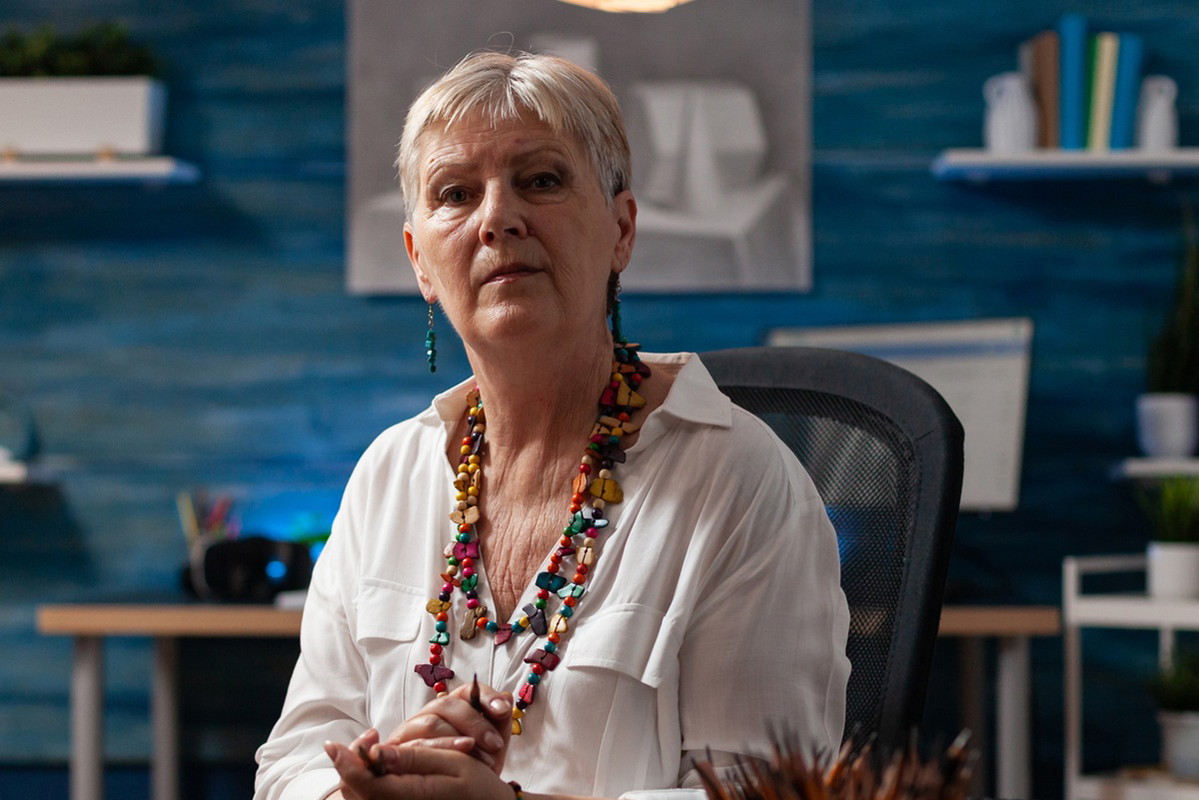 kobieta w wieku 55 plus z krótkimi włosami i koralami na szyi siedzi z ołówkiem w ręku smutna i zamyślona