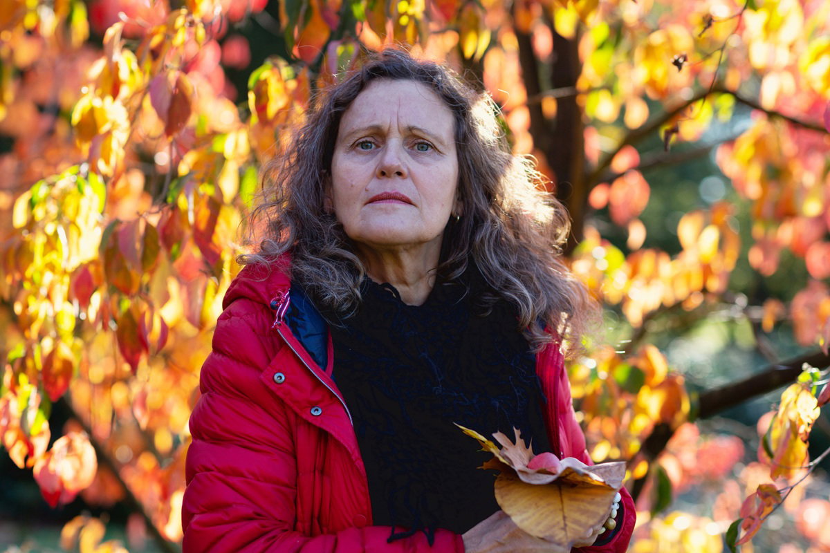 kobieta w wieku około 50 lat z długimi kręconymi włosami i w czerwonej kurtce stoi na tle jesiennych liści zaniepokojona