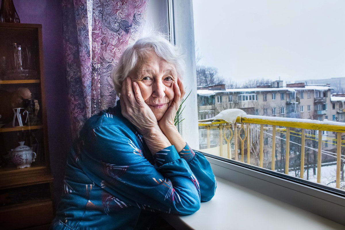 kobieta w wieku 70 plus w niebieskim stroju we wzory siedzi przy oknie zamyślona