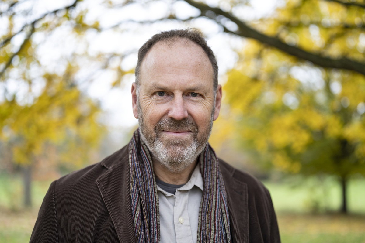 mężczyzna w wieku 55 plus z zarostem siwiejący w szaliku stoi na zewnątrz i lekko się uśmiecha