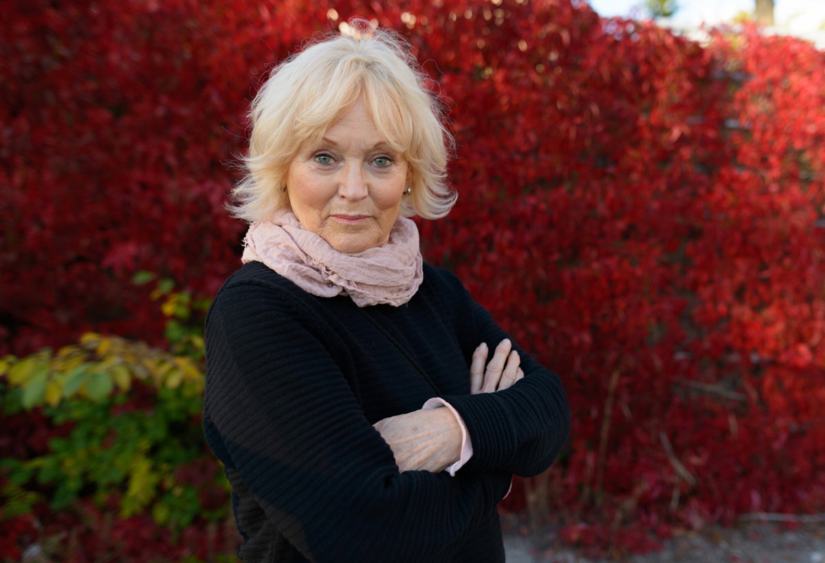 kobieta w wieku 60 plus blondynka w czarnym swetrze i pudrowo różowej apaszce stoi na zewnątrz uradowana