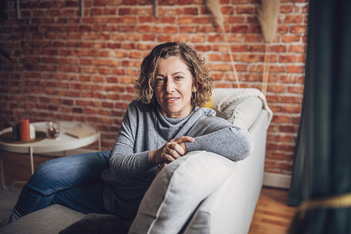 kobieta w wieku prawie 50 lat siedzi na kanapie w szarej bluzce zamyślona i lekko uśmiechnięta
