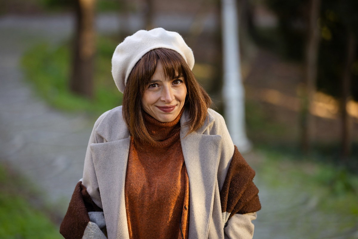 kobieta w wieku 30 plus w białym berecie uśmiecha się kpiąco stojąc na zewnątrz