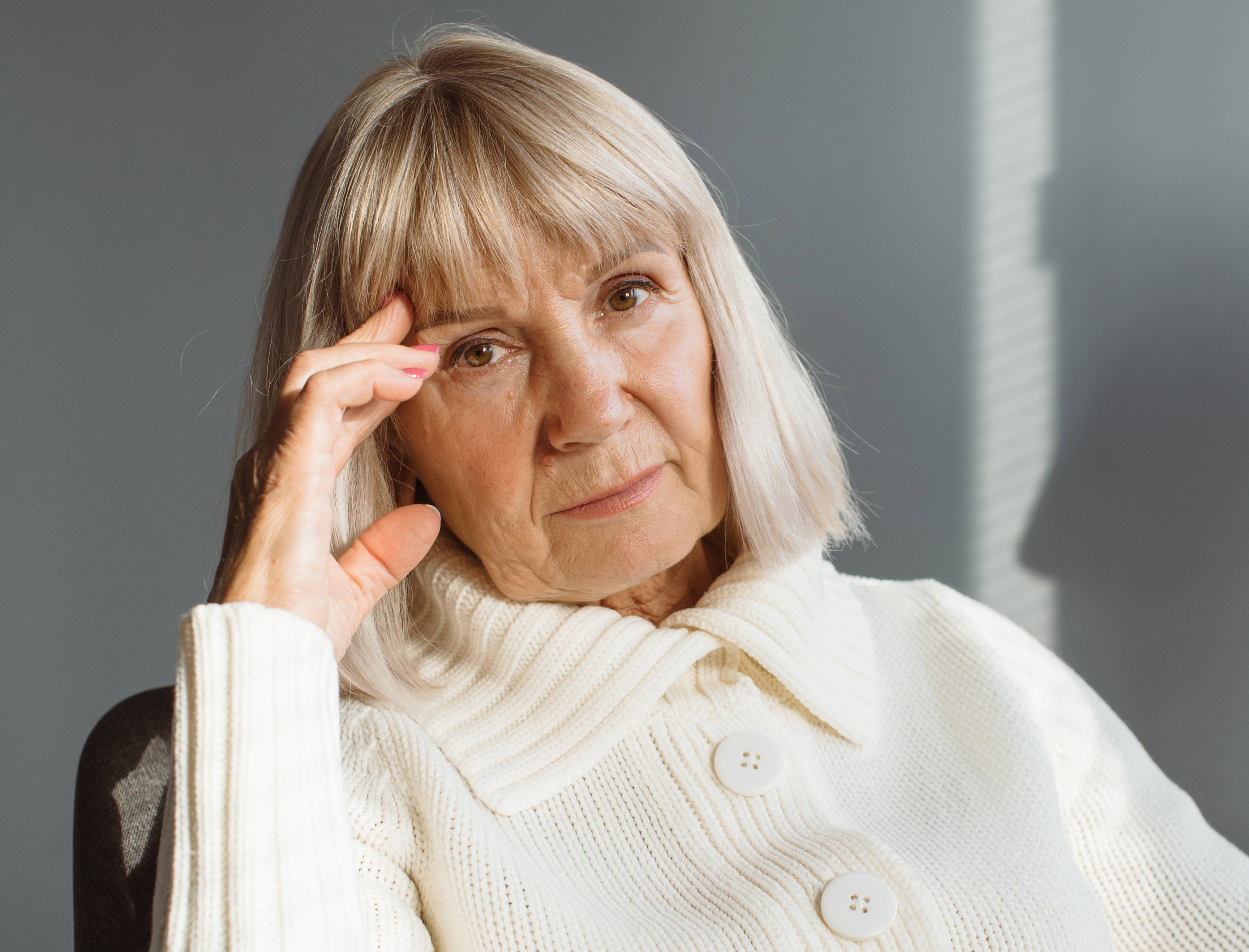 kobieta w wieku 55 plus blond włosy i w białym rozpinanym swetrze siedzi smutna i zamyślona