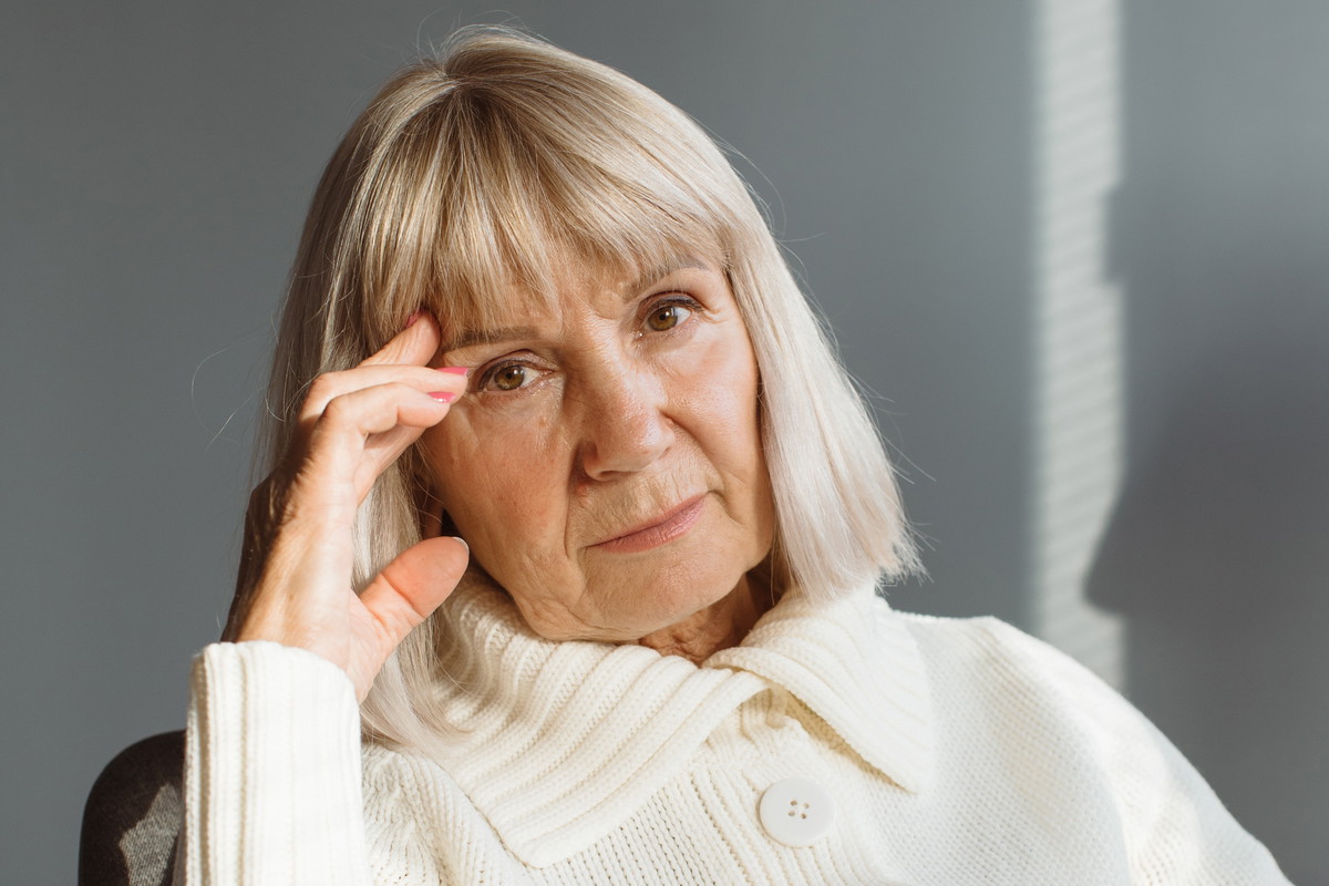 kobieta w wieku 55 plus blond włosy i w białym rozpinanym swetrze siedzi smutna i zamyślona