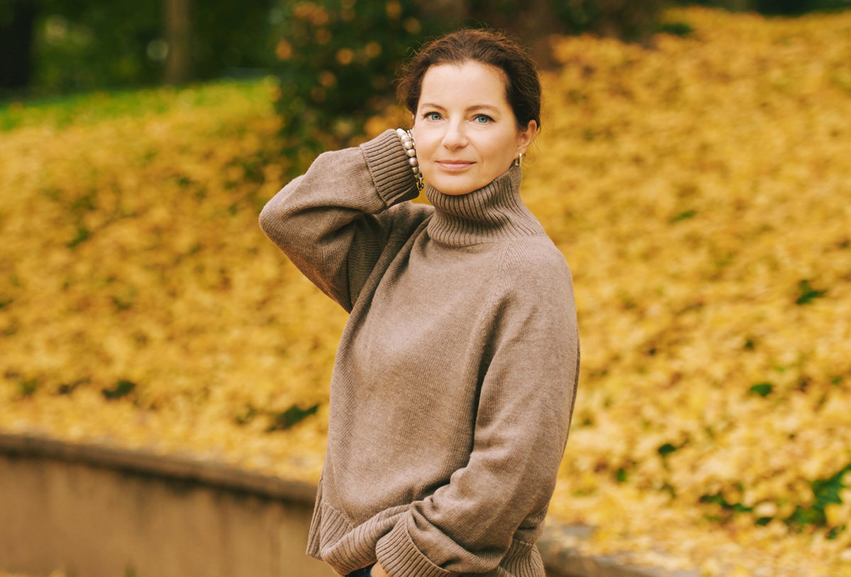 kobieta w wieku 35 plus stoi w jesiennym parku wyraźnie zrelaksowana i lekko uśmiechnięta