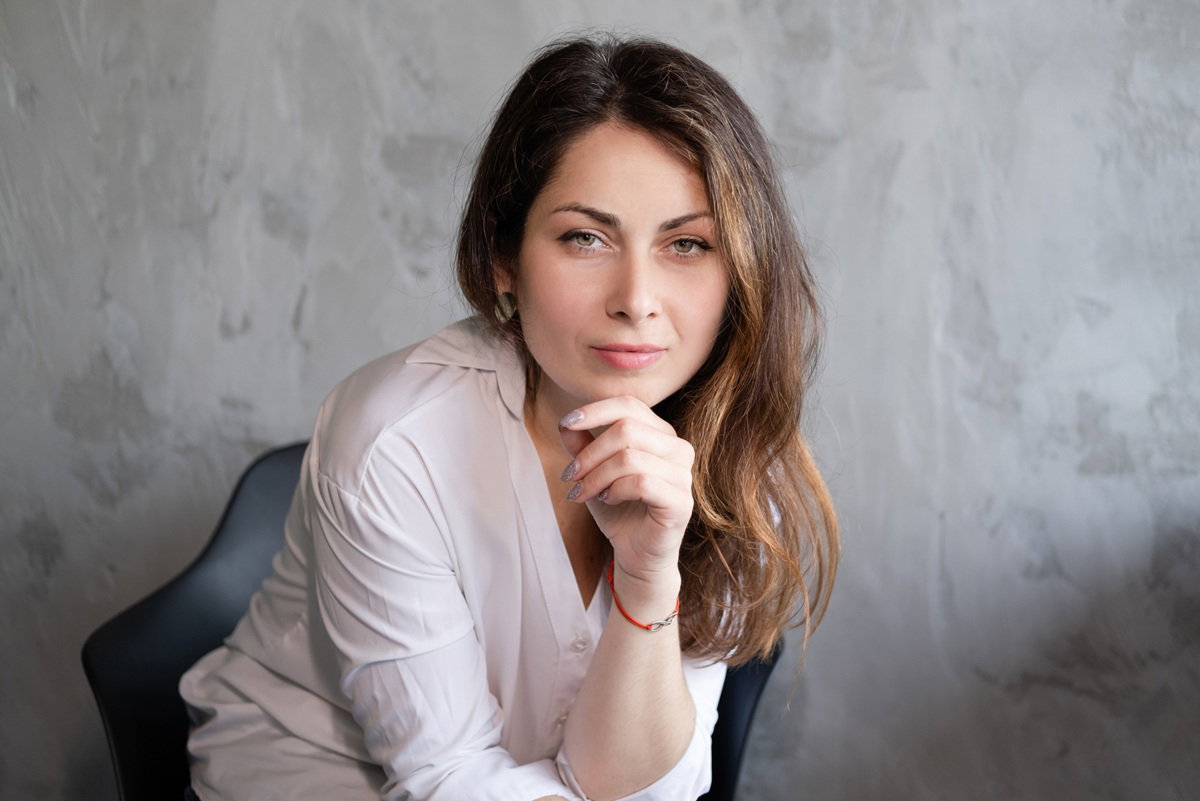 kobieta w wieku 30 plus siedzi z pewną siebie miną na fotelu na tle szarej ściany