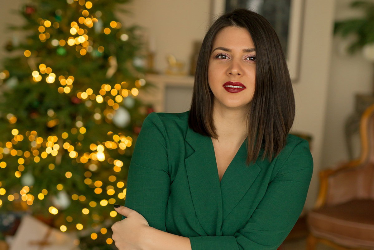 kobieta w wieku 30 plus w zielonej sukience stoi na tle choinki z kwaśnym wyrazem twarzy