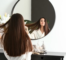 Beautiful young woman with healthy long hair looking in mirror at home
