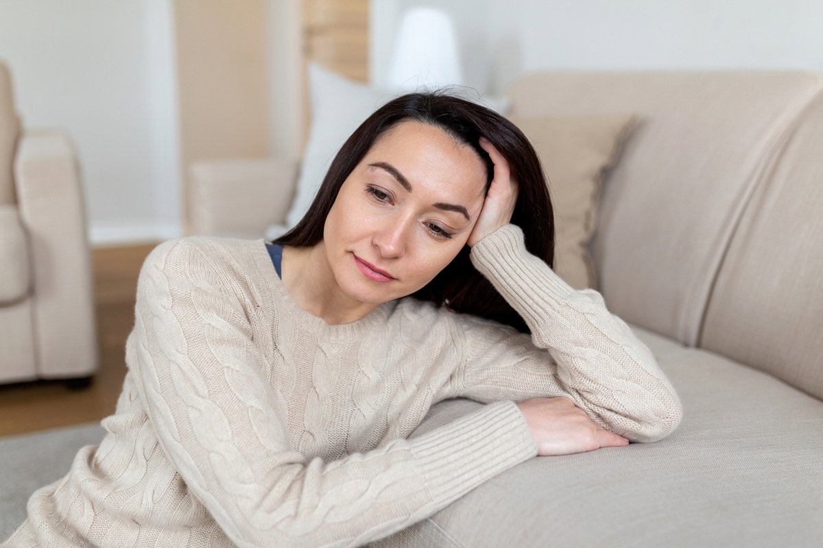 kobieta w wieku 35 plus w beżowym swetrze siedzi na podłodze obok kanapy zamyślona