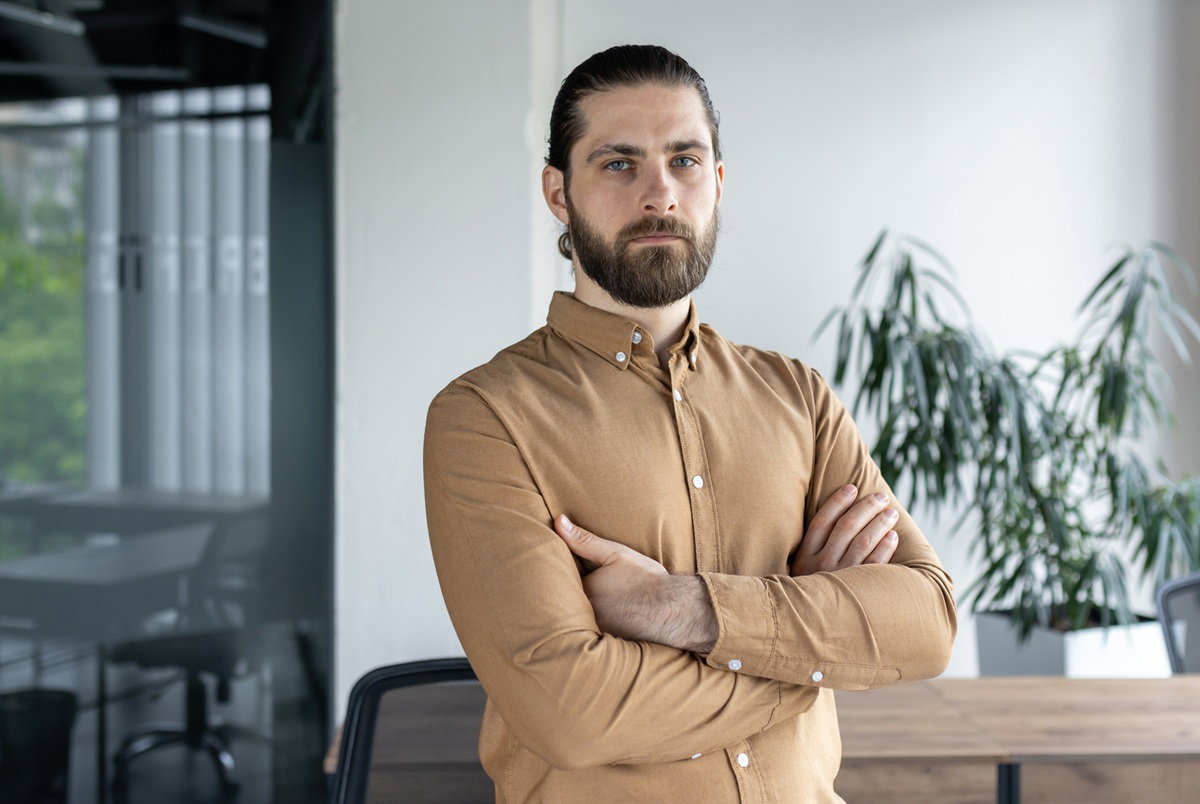 mężczyzna w wieku 40 plus z zarostem w musztardowej koszuli stoi w biurze zamyślony i zły