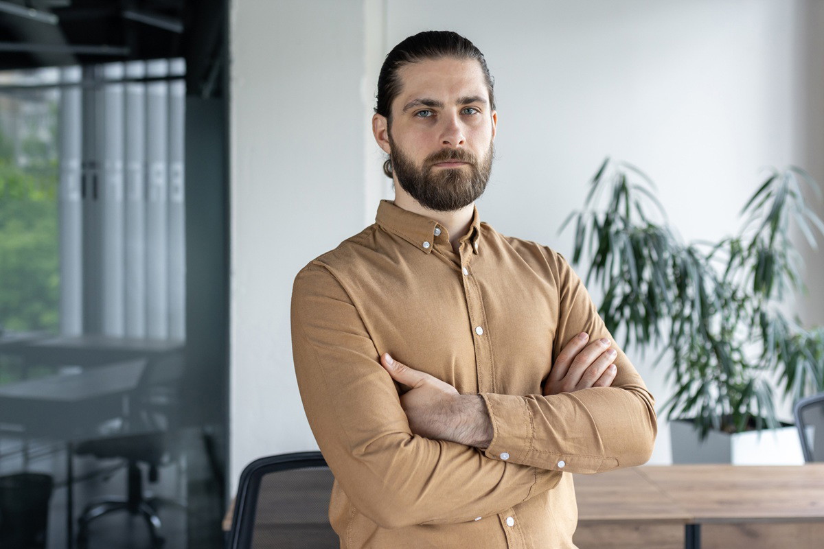 mężczyzna w wieku 40 plus z zarostem w musztardowej koszuli stoi w biurze zamyślony i zły