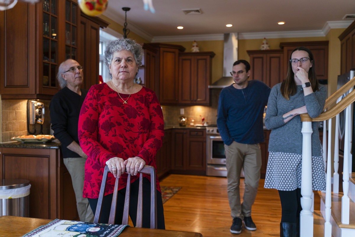 kobieta w wieku 60 plus w czerwonej bluzce w kwiaty stoi w kuchni za nią rodzina zamyśleni i poważni