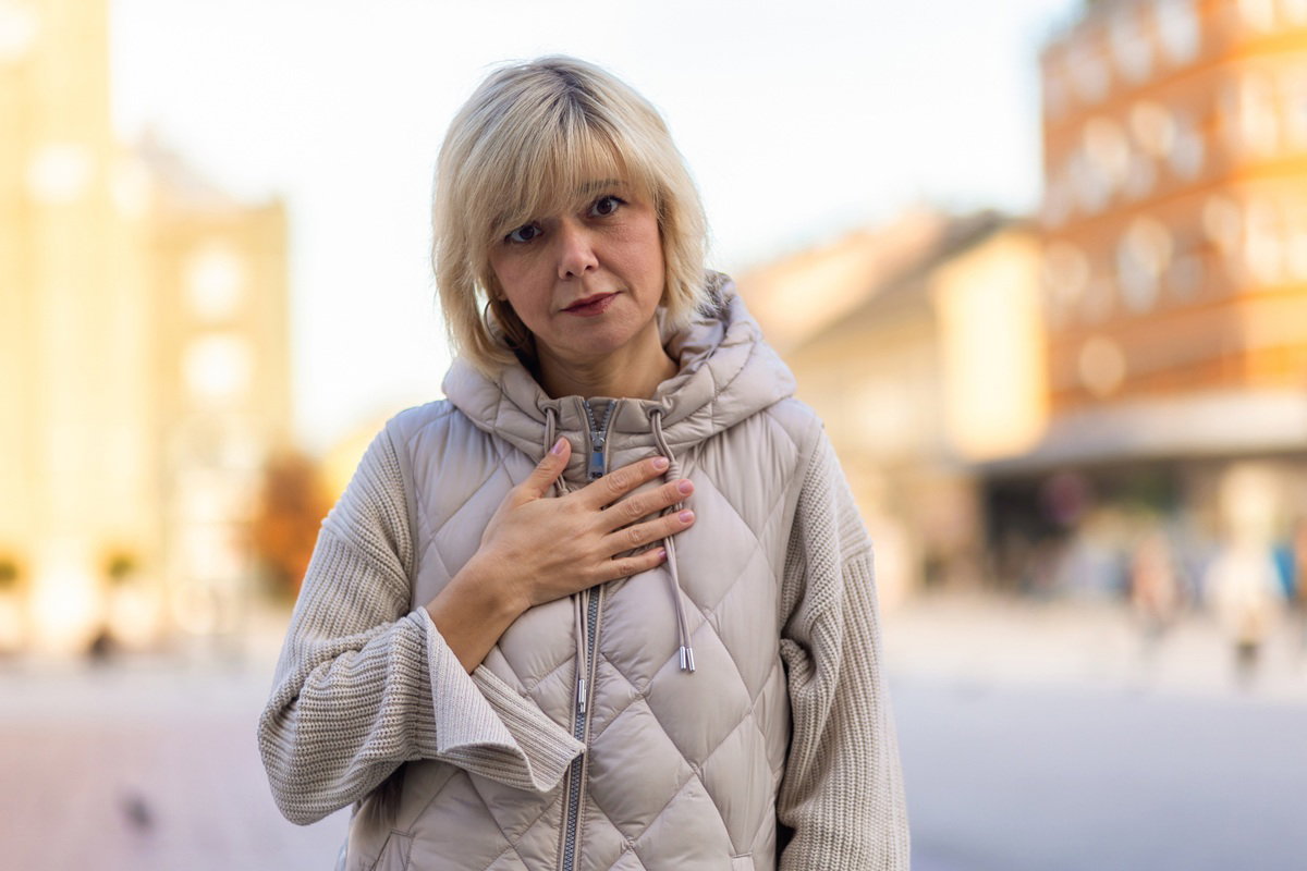 kobieta blondynka 40 plus w jasnym bezrękawniku i swetrze stoi na zewnątrz smutna i z poczuciem winy