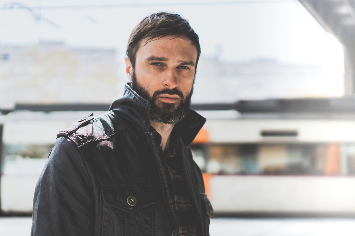 mężczyzna w wieku około 35 lat brodaty z wąsem w czarnej kurtce stoi na zewnątrz zmartwiony i smutny