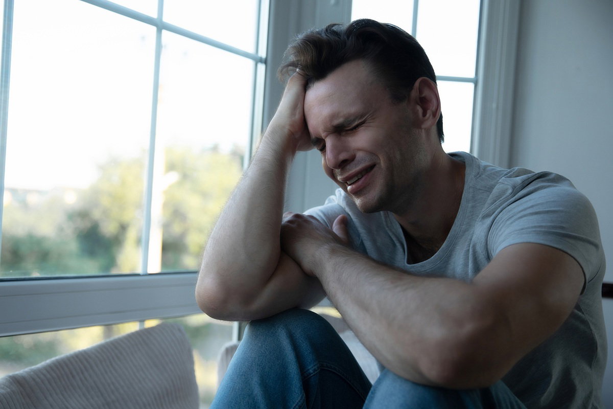 mężczyzna w wieku 20 plus siedzi przy oknie zrozpaczony i płacze