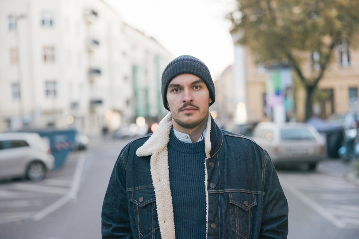 mężczyzna w wieku 35 plus z zarostem w jeansowej kurtce i w czapce stoi na ulicy smutny