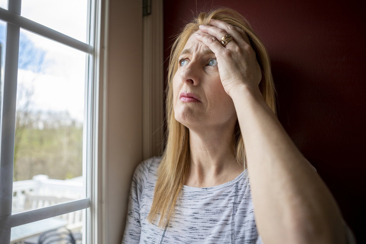 kobieta w wieku 45 plus z jasnymi włosami łapie się za głowę zmartwiona stojąc przy oknie