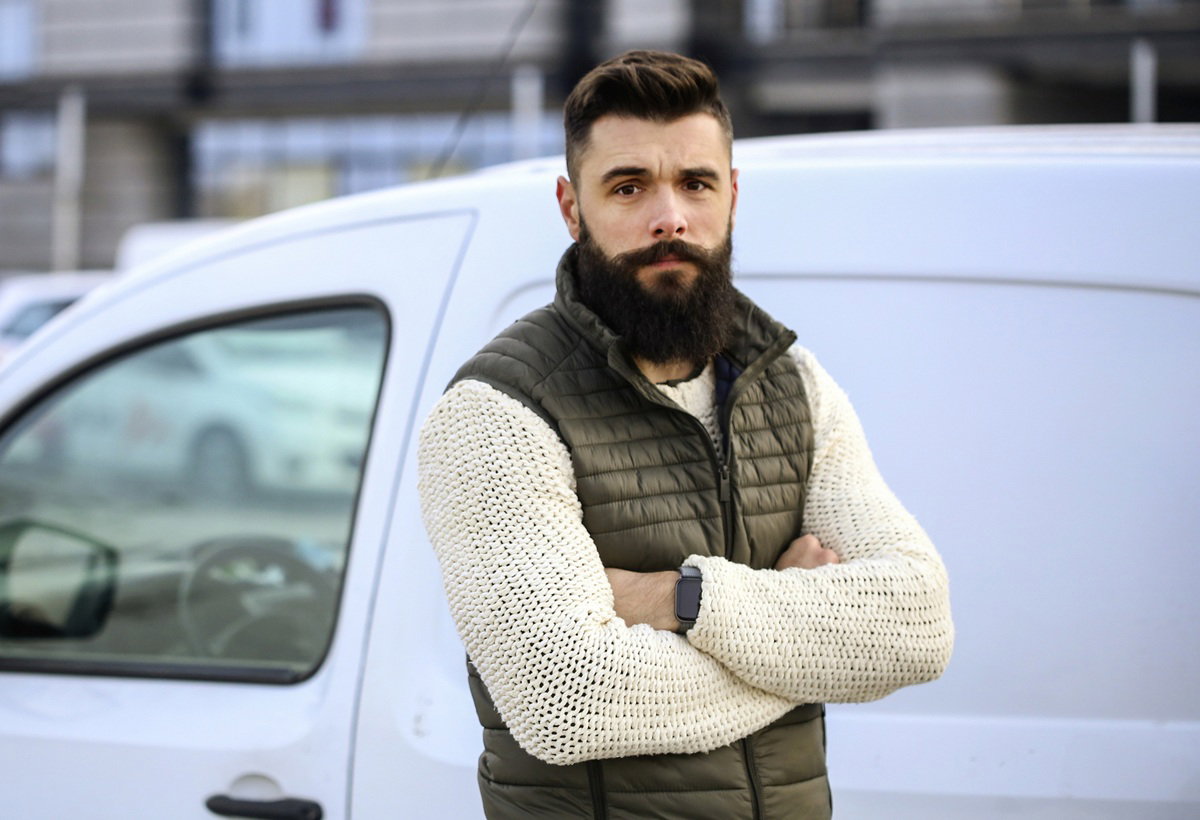 mężczyzna z brodą i wąsami w wieku 30 plus stoi na tle samochodu zmartwiony