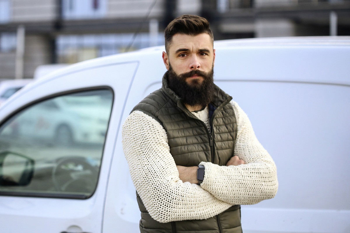 mężczyzna z brodą i wąsami w wieku 30 plus stoi na tle samochodu zmartwiony