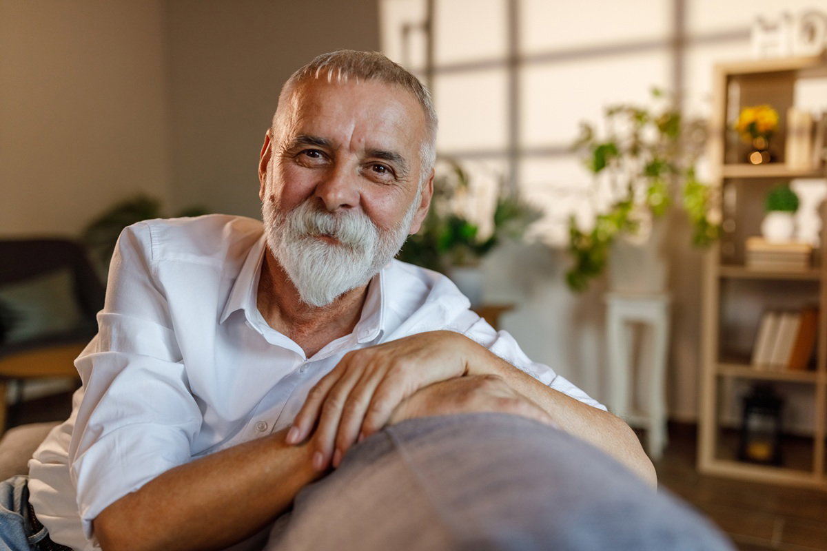 mężczyzna w wieku 60 plus w białej koszuli siwy z brodą i wąsami siedzi na kanapie i się uśmiecha
