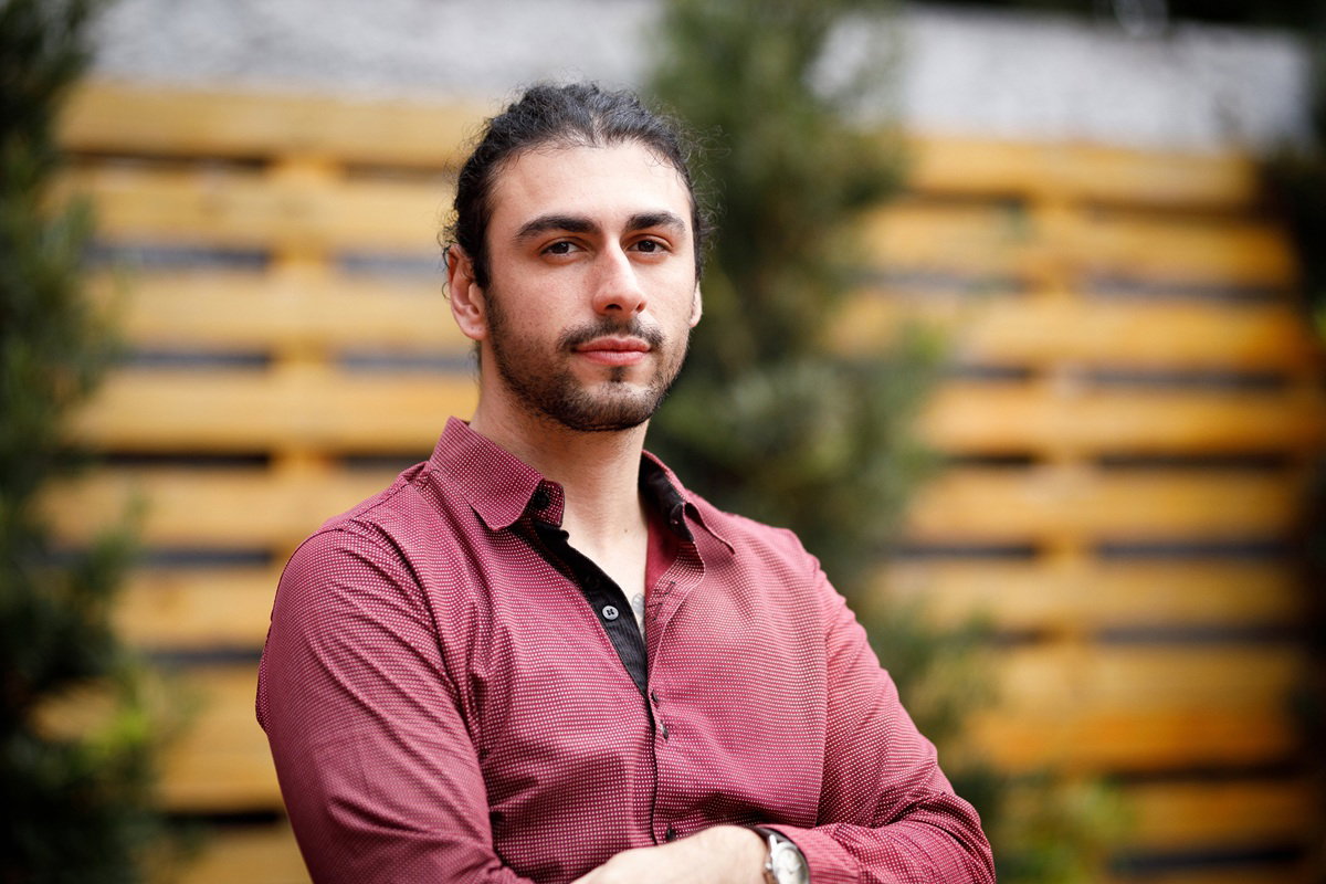 mężczyzna w wieku około 25 lat w czerwonej koszuli i z zarostem stoi na zewnątrz zamyślony