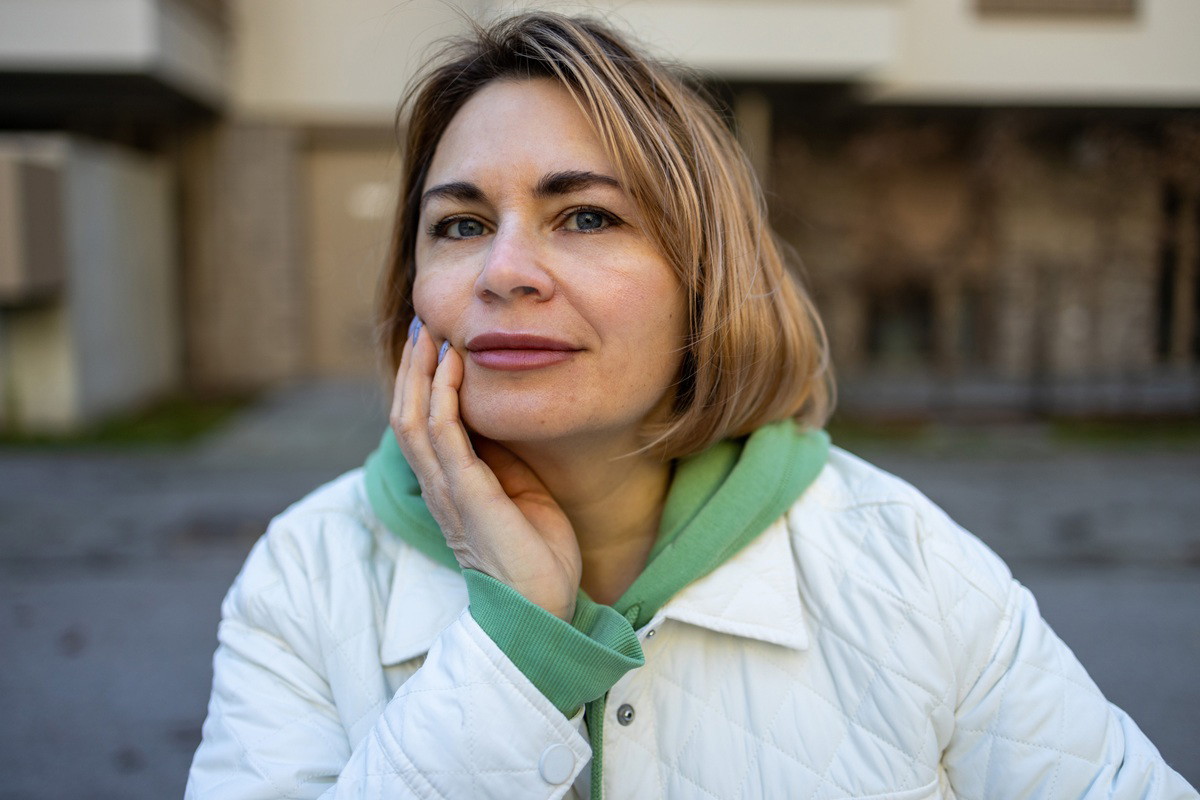 kobieta 45 plus blondynka w białej kurtce i zielonej bluzie stoi na zewnątrz zamyślona i z lekkim uśmiechem