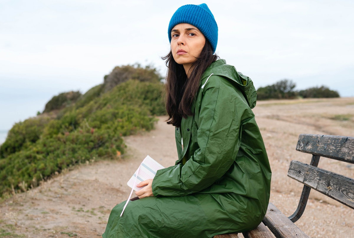 kobieta w zielonym deszczaku i niebieskiej czapce w wieku 30 plus siedzi załamana na ławce