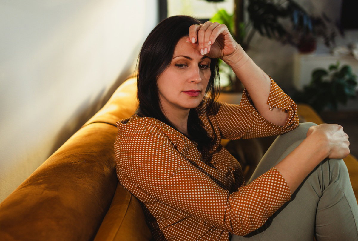 kobieta w wieku 40 plus z ciemnymi włosami w bluzce w groszki siedzi na kanapie zamyślona