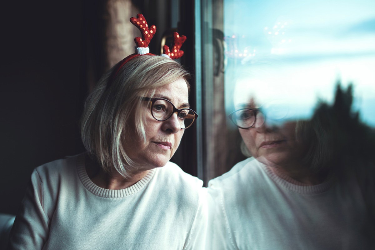 kobieta w wieku 60 plus w okularach i z różkami renifera na głowie patrzy w okno smutna