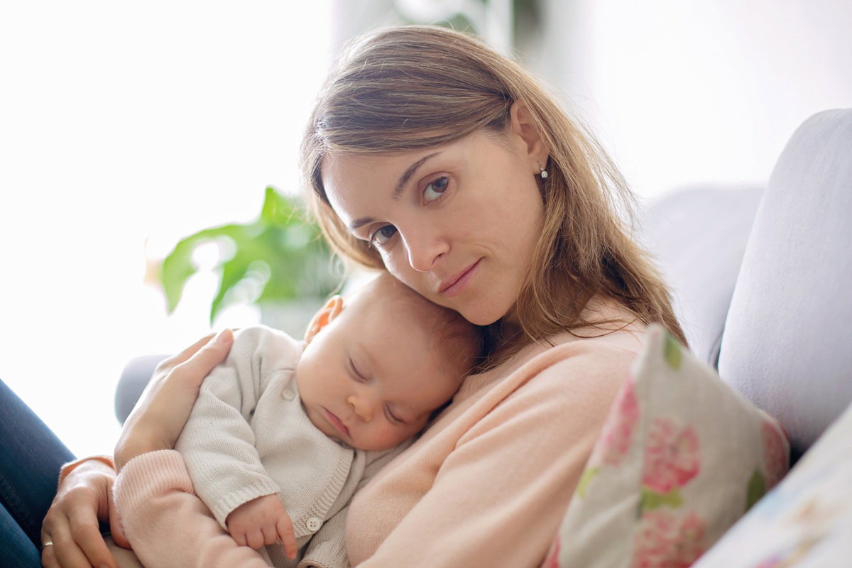 kobieta w wieku 30 plus z dzieckiem w ramionach siedzi na kanapie zamyślona