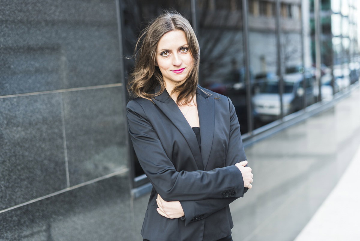 kobieta w wieku 30 plus stoi na tle biurowca z lekko kpiącą miną