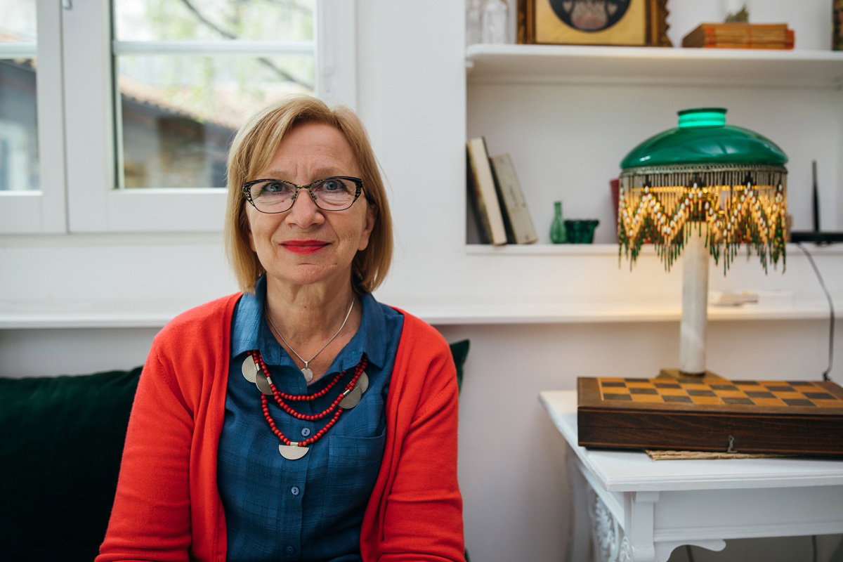 kobieta w wieku 50 plus w czerwonym sweterku siedzi przy oknie koło lampy z niepewnym spojrzeniem