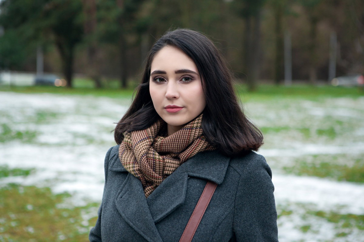 kobieta w wieku 30 plus z ciemnymi włosami i w płaszczu i kraciastym szalu stoi na zewnątrz zamyślona