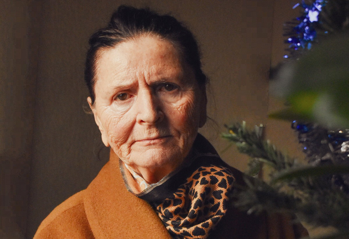 kobieta w wieku 70 plus w pomarańczowym płaszczu z apaszką w panterkę stoi przy choince zasmucona