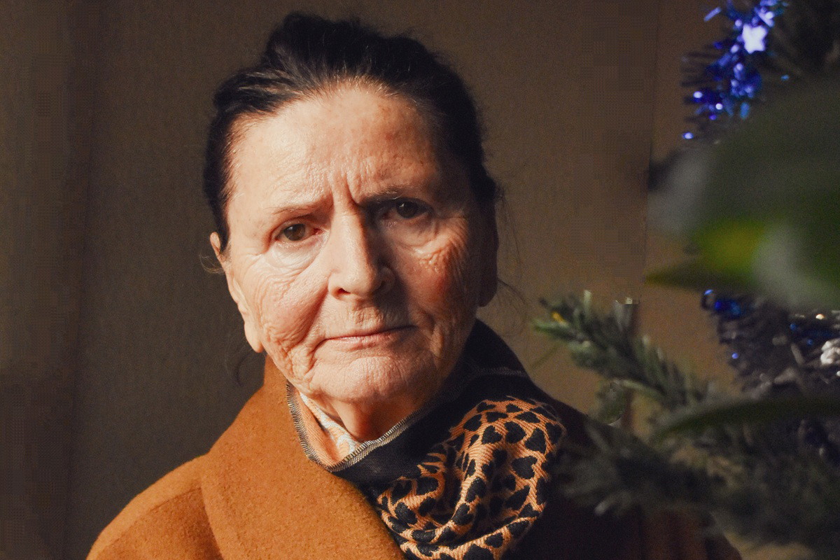 kobieta w wieku 70 plus w pomarańczowym płaszczu z apaszką w panterkę stoi przy choince zasmucona