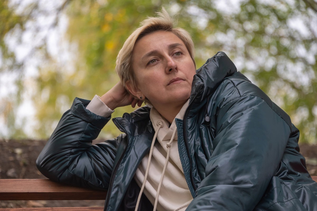 kobieta w wieku około 40 lat w zielonej kurtce siedzi na ławce w parku zamyślona