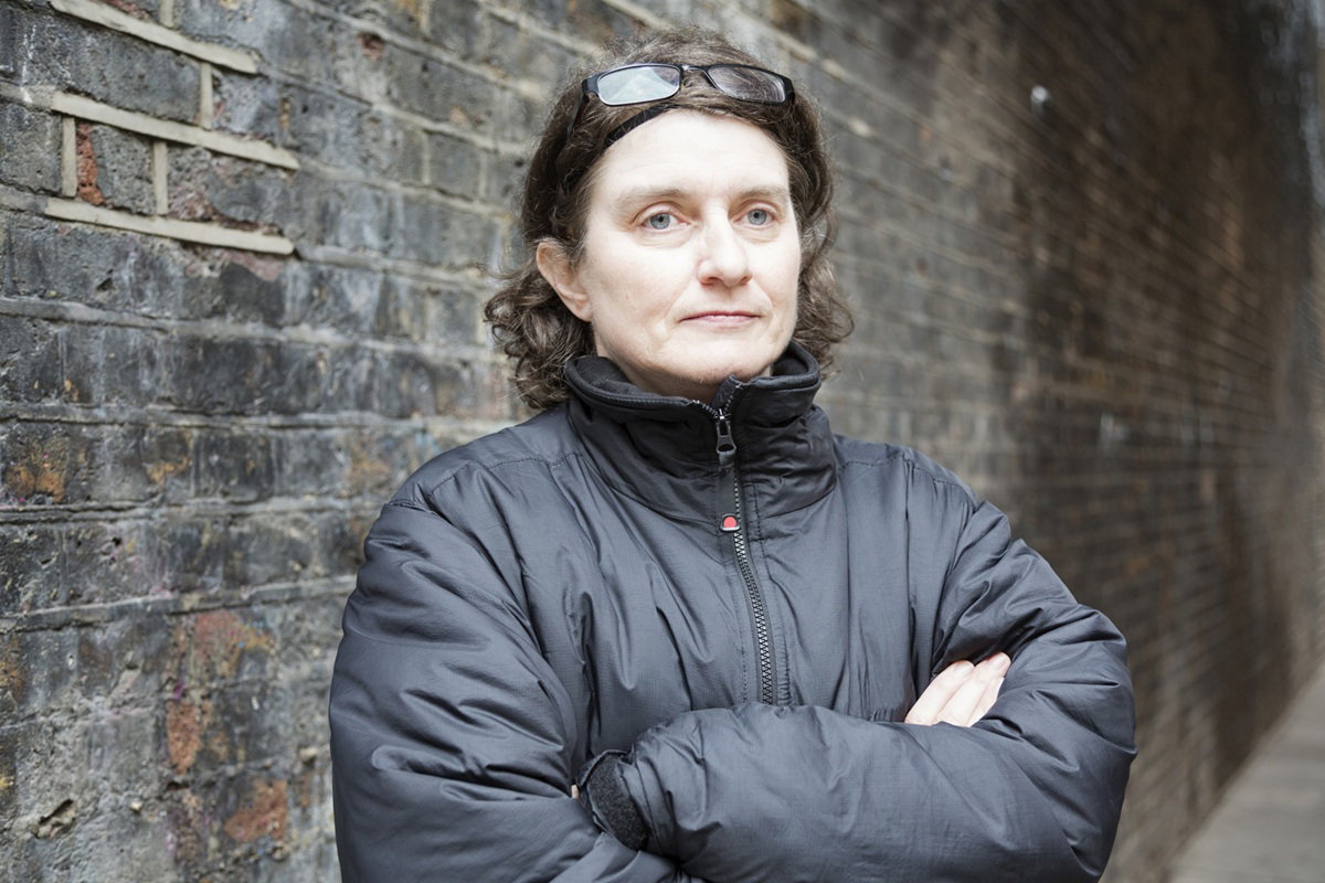 kobieta w wieku 50 plus w czarnej kurtce i w okularach na głowie stoi na tle muru i patrzy w dal