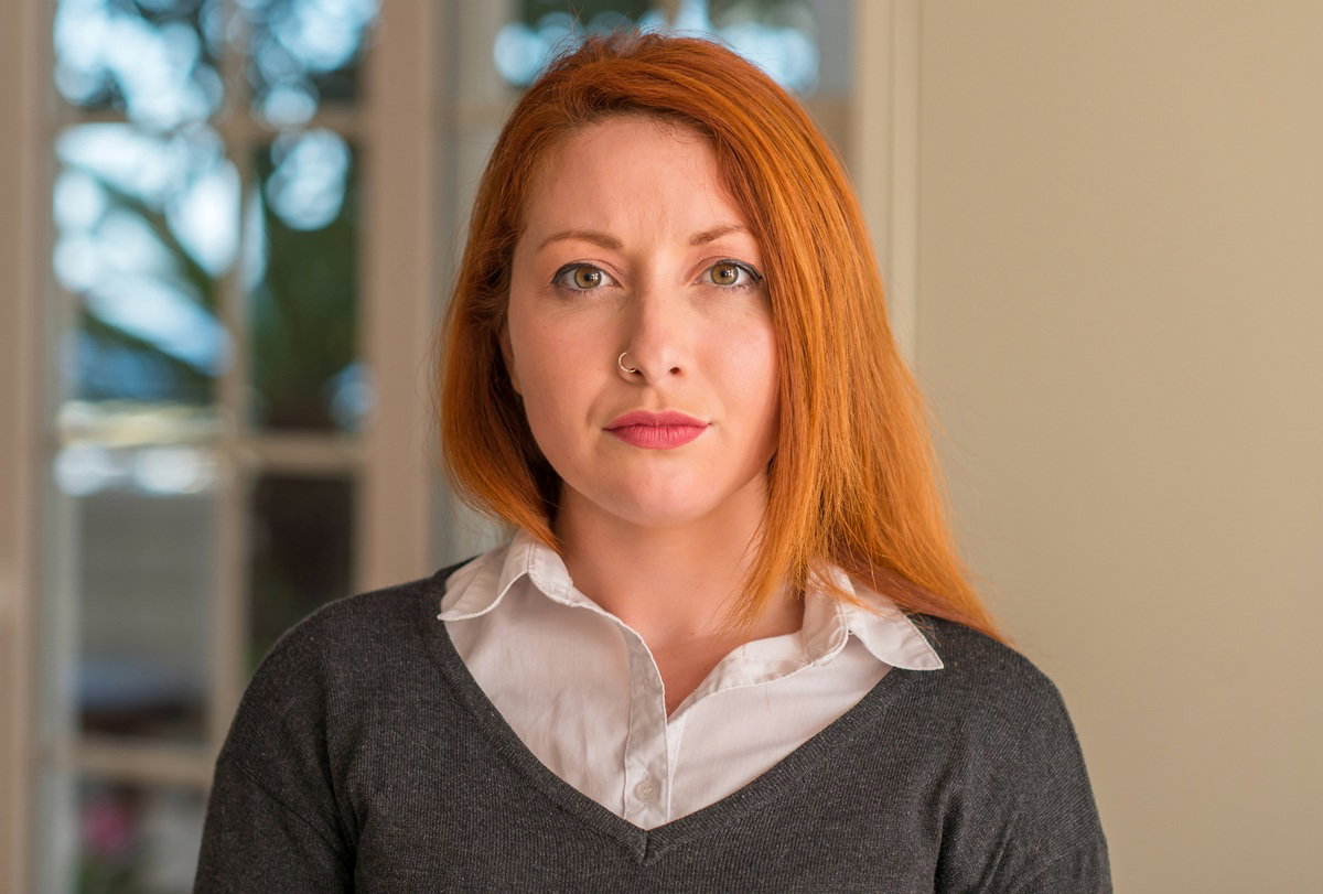 kobieta w wieku 30 plus z rudymi włosami stoi na tle okna poważna i zamyślona