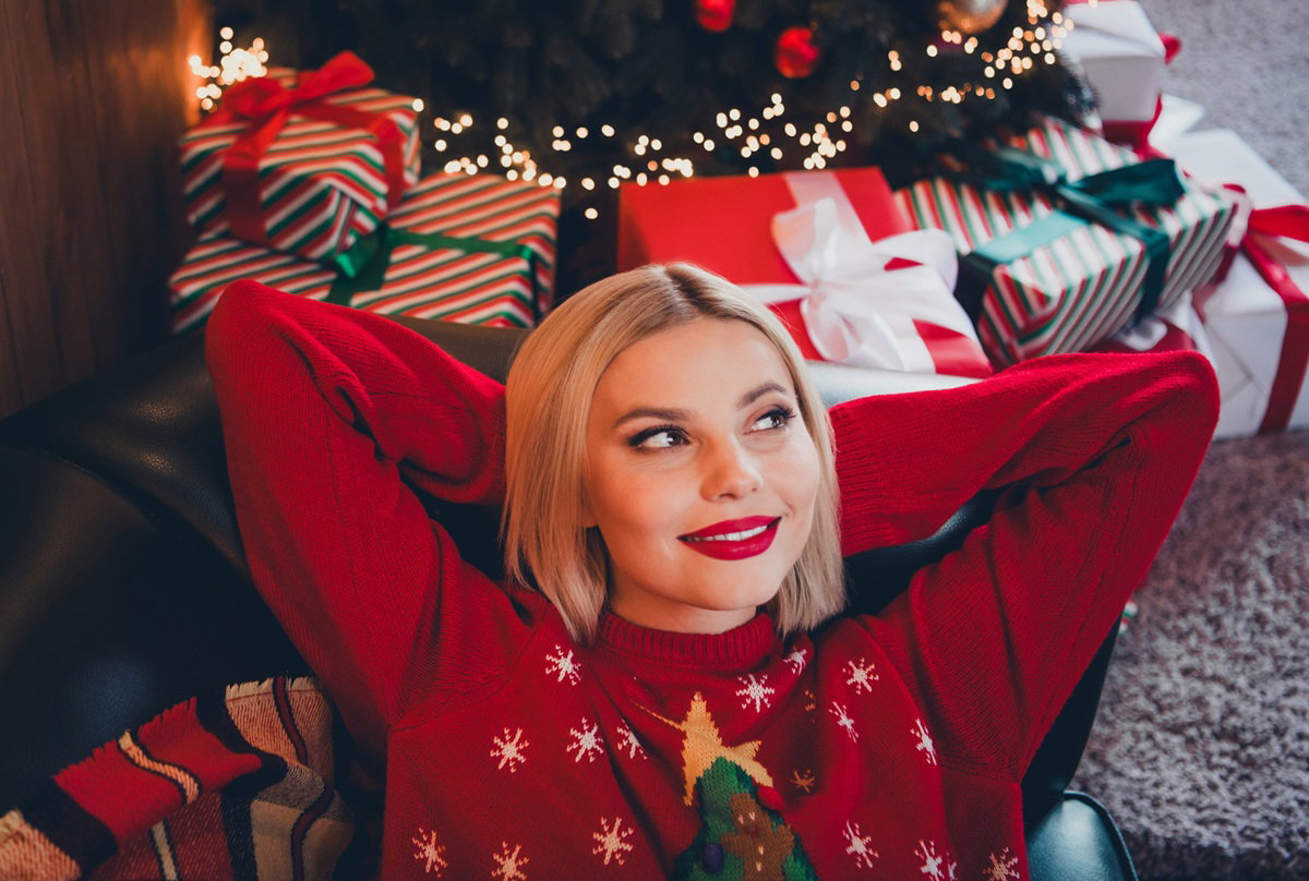 kobieta w wieku 30 plus blondynka w świątecznym swetrze leży koło choinki zadowolona