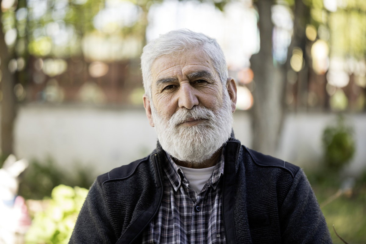 mężczyzna w wieku 55 plus siwy z brodą i wąsami siedzi na zewnątrz z poważnym wyrazem twarzy