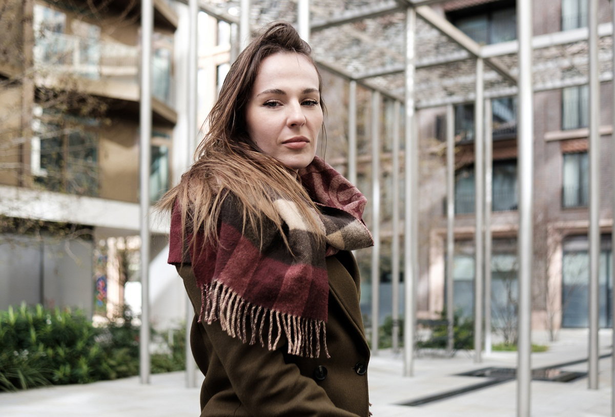 kobieta w wieku 30 plus z długimi włosami stoi na tle nowoczesnego budynku z poważnym wyrazem twarzy