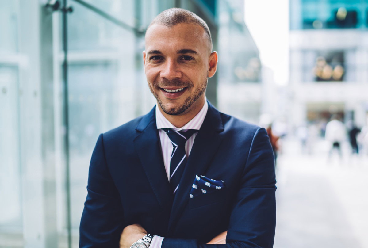 mężczyzna w wieku 35 plus w granatowej marynarce stoi na tle biurowca i lekko się uśmiecha