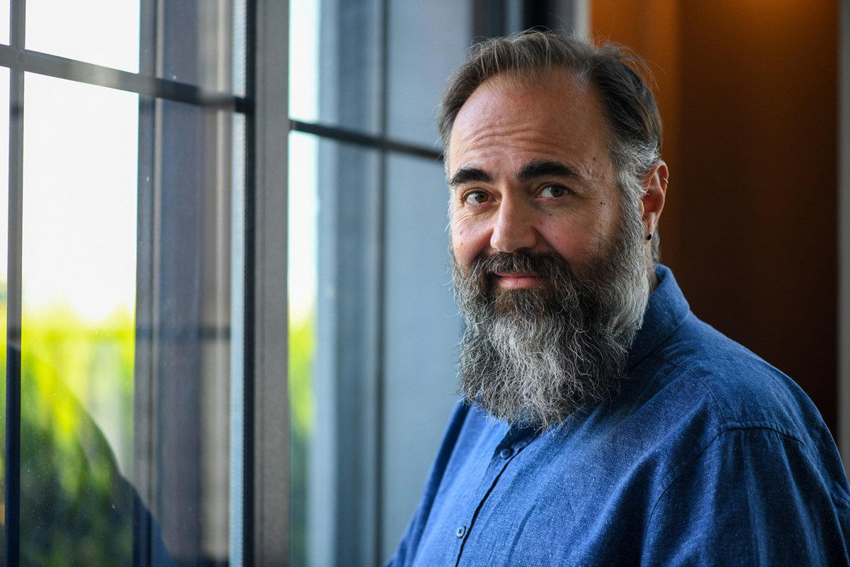 mężczyzna z wąsami i brodą łysiejący i siwiejący w wieku 45 plus stoi przy oknie z naiwnym spojrzeniem