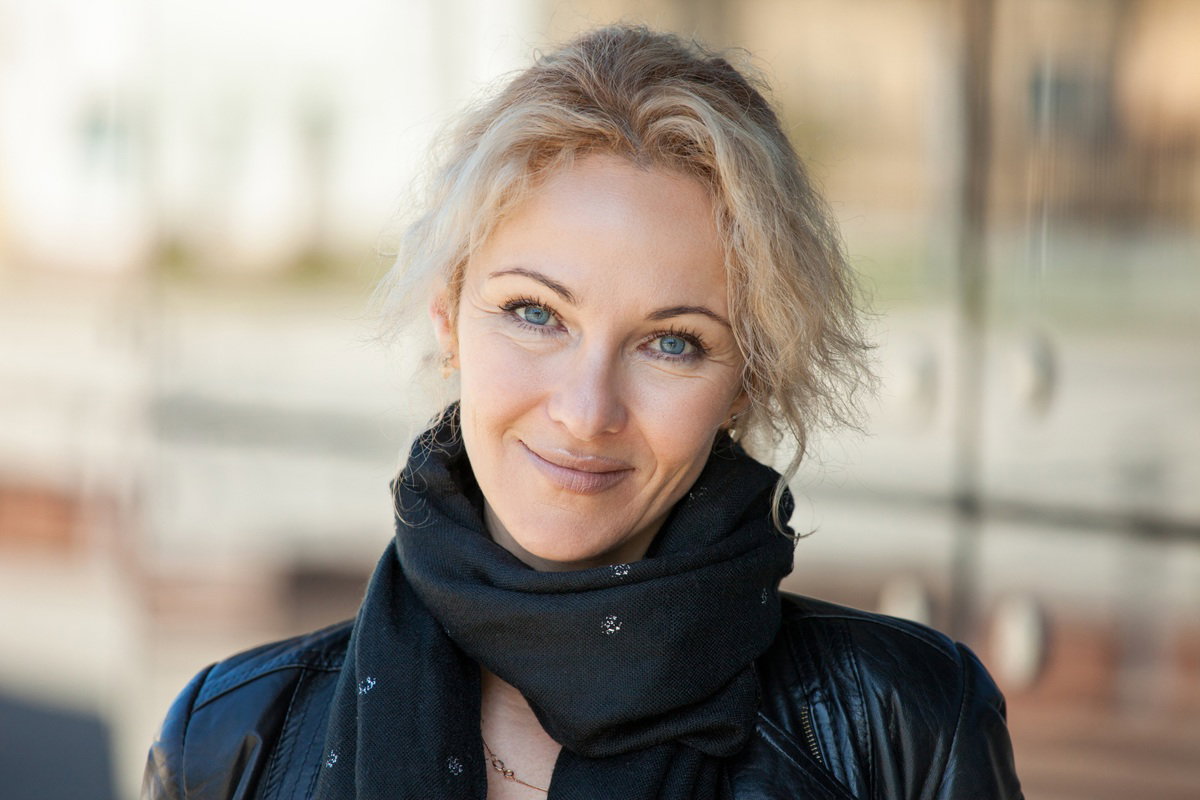 kobieta w wieku 40 plus blondynka z szalem na szyi i w skórzanej kurtce stoi na ulicy i się uśmiecha