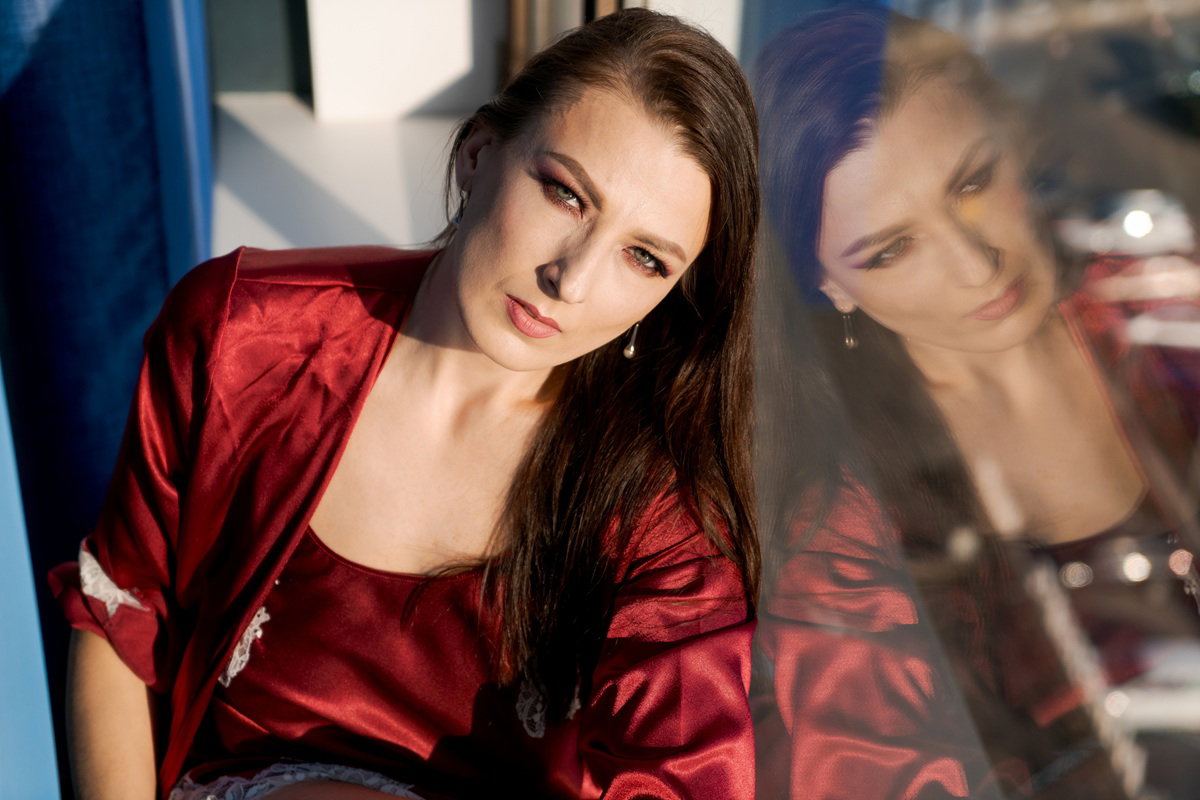 kobieta z długimi włosami 35 plus w szlafroku i w koszulce do spania siedzi przy oknie rozmarzona