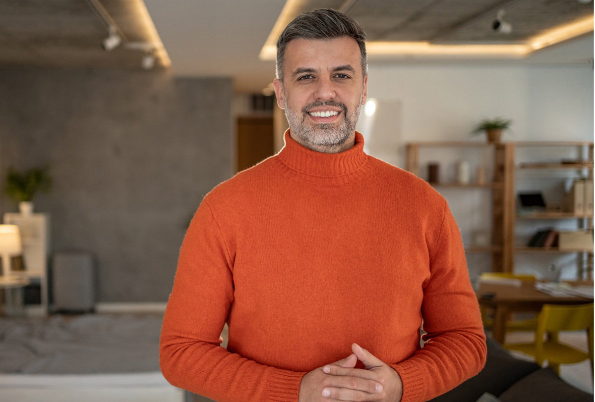 mężczyzna w wieku 35 plus w jasnoczerwonym swetrze z golfem stoi w salonie ze splecionymi dłońmi i uśmiecha się