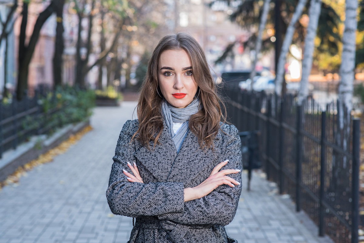 młoda kobieta w wieku 25 plus stoi na ulicy w płaszczu w jodełkę z lekko zdziwionym wyrazem twarzy